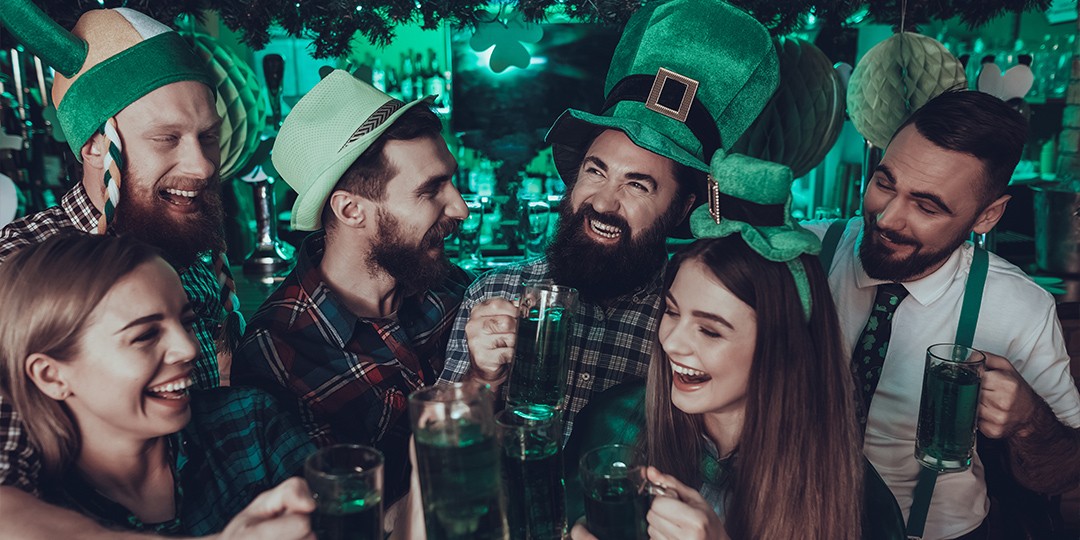 A group of people in St Patrick’s Day-themed costumes, cheersing green drinks and laughing. 