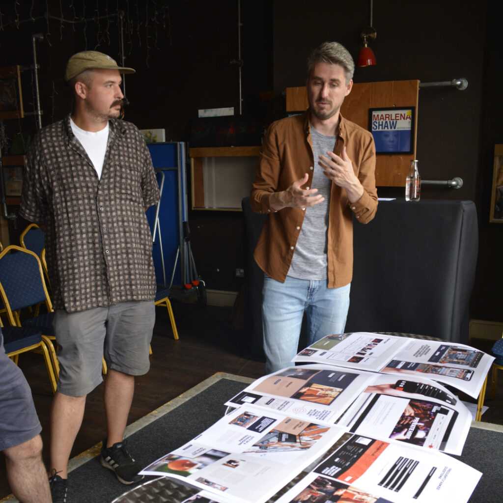 A photograph of Scott Barker, lead designer at Brew, and the team from 1000 Trades in a workshop discussing the new website. Mood boards and website designs are scattered on a table in front of them.